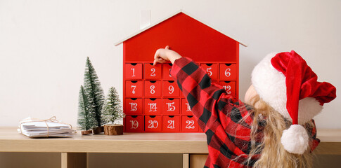 Little girl in Santa hat near wooden shelf unit with Christmas advent calendar and letters against light wall