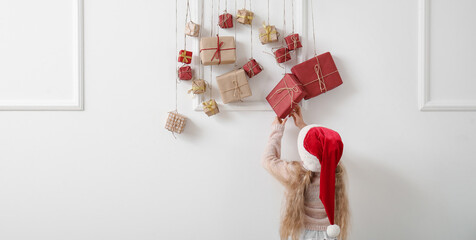 Cute little girl in Santa hat and Christmas advent calendar hanging on light wall
