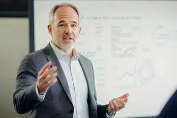 A middle-aged Caucasian man confidently leading a business presentation before a whiteboard, gesturing towards the data. His commanding presence reflects his authority and knowledge.