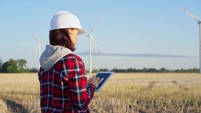 Woman engineer wearing a white protective helmet is taking notes with a tablet computer on a field with wind turbines, as the sun sets. Clean energy and engineering concept