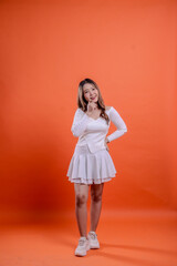 Full length beautiful Asian female in white top standing cheerful hands holding waist and chin isolated orange background. for the concept of fashion, beauty, advertising,