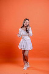 full length beautiful woman in white top standing singing cheerfully to camera holding mic and looking forward, voice recorder, orange background isolation. for fashion, advertising, entertainment