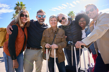 Portrait group of mature tourist people posing hugging happy and looking at camera celebrating together journeys. Diverse friends of tourism with luggage on street on sunny winter day
