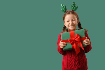 Cute little girl in reindeer horns with Christmas gift box on green background