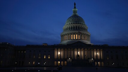 Showing the East side of the US Capitol with lights and flags waving in Washington DC at night set against a blue purple sky.