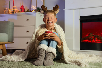 Little boy in reindeer horns with cup of cocoa sitting near fireplace at home on Christmas eve