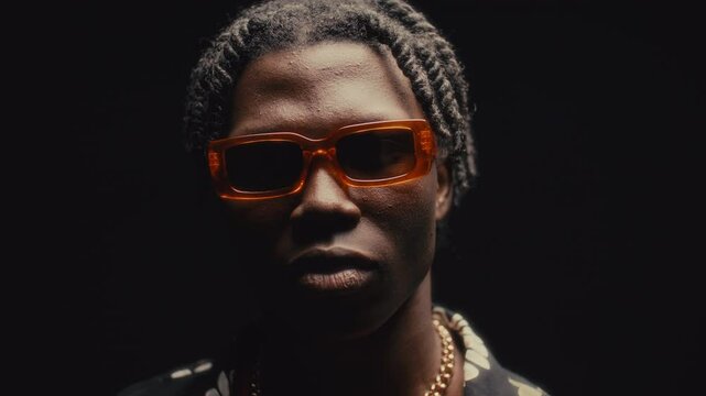 Young Black man with dreadlocks wearing bright orange sunglasses, chain necklace and patterned shirt looking at camera with confident expression against dark background in studio. Video portrait
