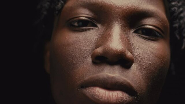 Young African American man with braids looking directly to camera, then gazing up pensively against dark background in studio. Video portrait, close-up view