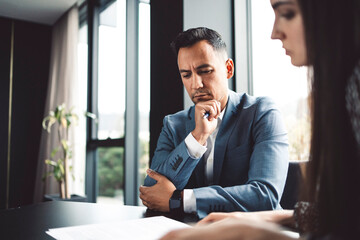 Two business people in business clothes are sitting in an office looking at a contract. One person is giving advice to the other. After signing the contract, they shake hands. Together they look out o