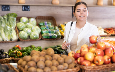 Portrait of positive interested young woman buying fruits in greengrocery store, choosing ripe...