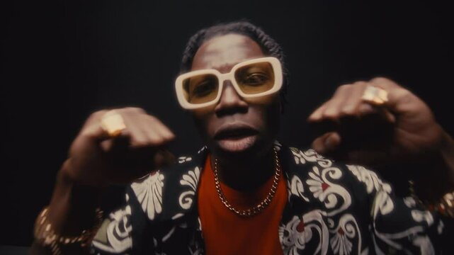 Young Black hip-hop musician in sunglasses, gold chain and rings and stylish outfit gesturing as rapping on camera with expression. Dynamic zoom, studio shot