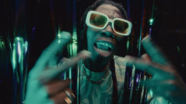 Stylish Black rapper in oversized white glasses and chain necklace performing intense rap to camera against shimmering tinsel curtain illuminated with multicolored lights