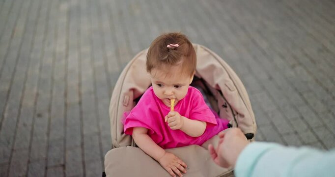Little girl in pink dress nibbles breadstick and sits in stroller during rest and walk with her mother in city