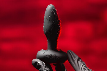 Silicone anal plug in a female hand in black latex gloves on a black background. Massager with...
