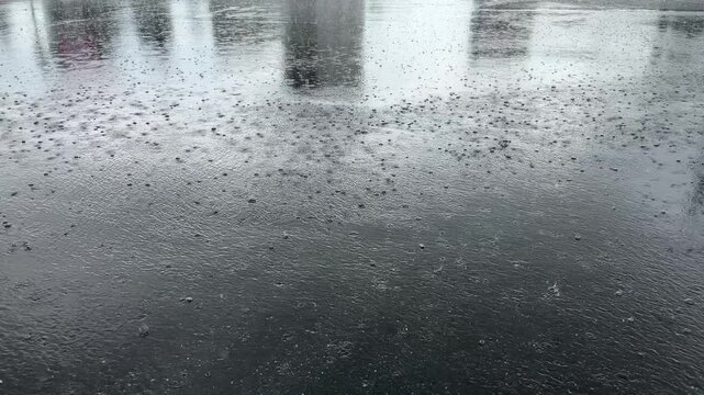Drops of rain pelt the asphalt, creating a symphony of splashes as the water collects in glistening puddles on the gray surface.