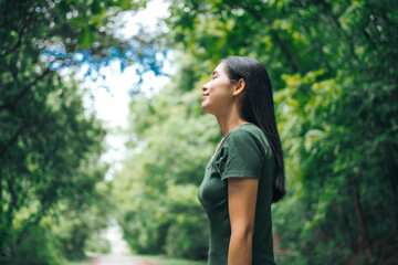 An Asian woman is standing happily in a green forest park, enjoying the fresh air and nature. She...