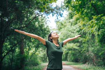 An Asian woman is standing happily in a green forest park, enjoying the fresh air and nature. She takes a deep breath, looks up at the morning sunlight, and feels relaxed and peaceful.