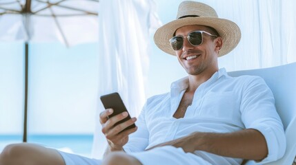 Smiling man in a straw hat and sunglasses using his smartphone at the beach, enjoying a sunny day and relaxing by the ocean