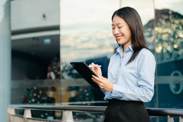 Obraz premium Asian woman with a smile standing holding notepad and tablet at out side office. big city on business center