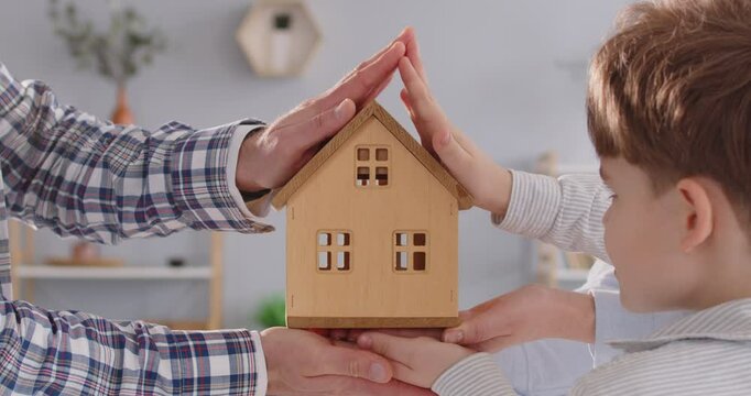 Family with child creates home symbol from hands over wooden model of house. Hands of parents and child are united over model of house, symbolizing unification of family and dream of owning home.