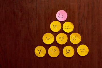 Pink Smiley Among Yellow Unhappy Faces Pyramid on Wooden Background