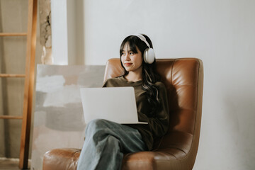 Focused Work, Serene Sounds: A young woman, immersed in her work, sits comfortably in a leather armchair, headphones on, laptop open, lost in the flow of her creative process.