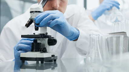 Fototapeta premium Young scientist or researcher in protective workwear studying new virus in microscope to develop antidote for fighting coronavirus