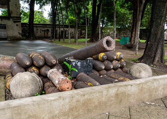 An old cannon rests atop a pile of mortar shells, all arranged in a concrete planter in a park setting.