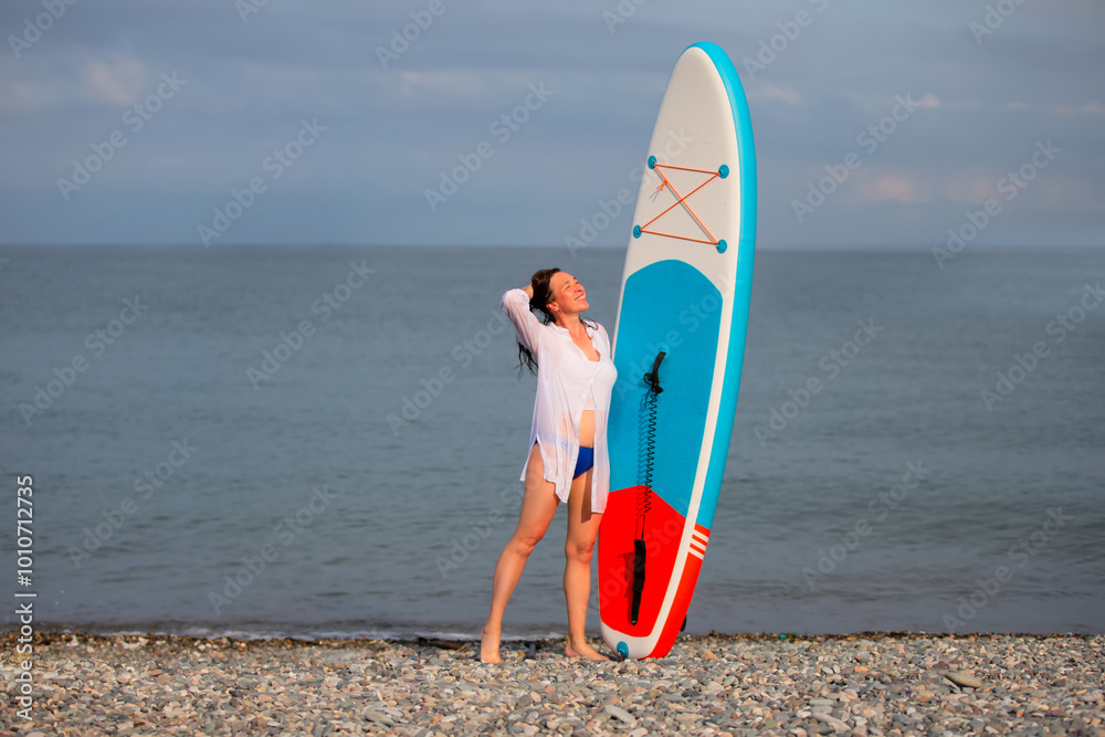 Wall mural a woman stands on the beach with a sup board. she is wearing a white shirt and blue swimming trunks.