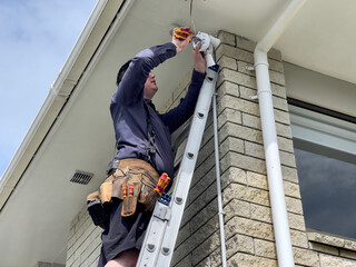 Caucasian man wearing a leather tool belt, holding pliers while standing on a ladder installing a security sensor light to the underside of a soffit on the corner of a brick clad house in New Zealand © Harriet
