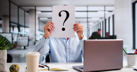 Businessman Holding Paper With Question Mark In Front Of Face At Desk
