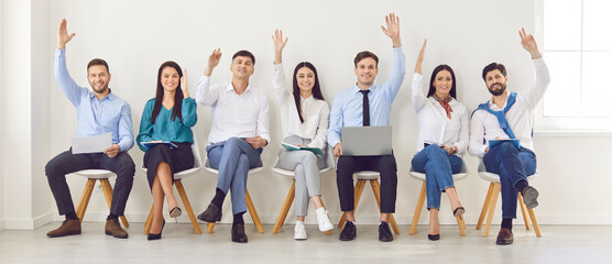 Young cheerful colleagues and company employees raising hands to vote at conference sitting on chairs in row in meeting room. Smiling business people men and women voting in office during a meeting