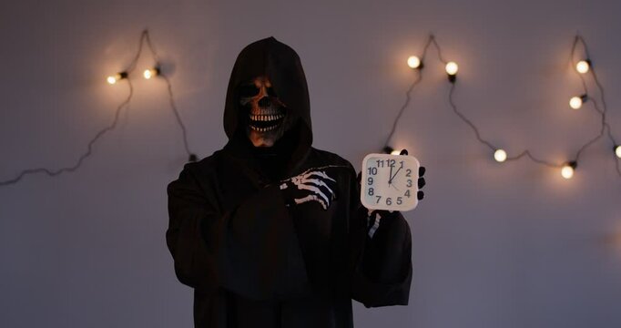 Death reminding that time is ticking away. Person dressed in a dark hooded robe of the Grim Reaper pointing a finger at a clock in hand as a metaphor for life's countdown. Life and death concept