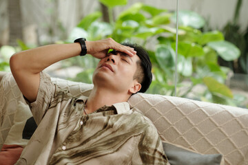 Young man lying on sofa and holding his head with hand, he having headache