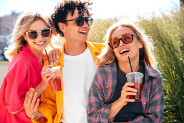 Young three stylish friends posing in the street. Fashion man and two cute female dressed in casual summer clothes. Smiling models having fun. Cheerful women and guy outdoors, Hold and drink lemonade