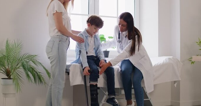 Doctor traumatologist sitting on couch in exam room with child boy examining his injured broken leg in cast in clinic. Pediatrician or family doctor consulting patient. 4k video. Slow motion video