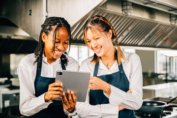 Two renowned chefs discuss video blog in a professional restaurant kitchen using a tablet. Professionals smiling searching reading and communicating with modern technology.