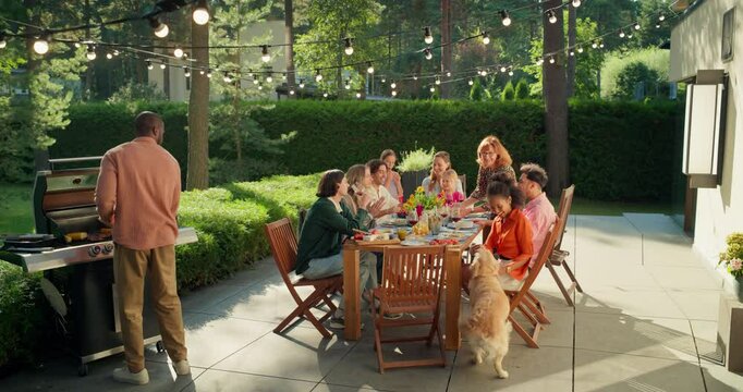 Lively Garden Party with Friends and Family Enjoying Food, Drinks, and Laughter Under String Lights Outdoors. Black Man Bringing Food From the Grill while Others Toast and Enjoy Joyful Atmosphere