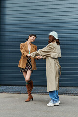 Two women dressed in trendy outfits smile and hold hands in front of a blue metal garage door