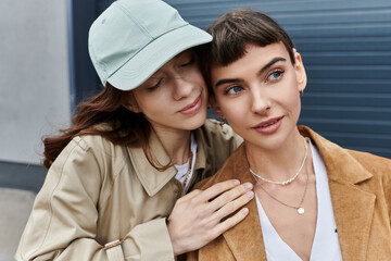 Two young women, dressed in trendy attire, share a tender moment outdoors, surrounded by the urban...