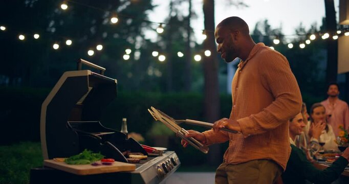 African Man Grilling a Beef Patty on a Barbecue in a Backyard with Friends Enjoying Drinks and Conversation in the Background. Young Black Man Dancing, Enjoying the Party Atmosphere
