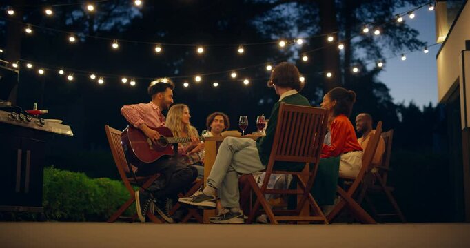 Friends Gather Around a Table in a Home Backyard, Smiling and Singing Along as One Friend Plays a Guitar. Evening Scene is Filled with Laughter, Music and a Warm Sense of Togetherness
