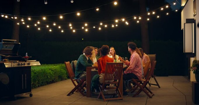 Diverse Group of Friends Enjoys an Outdoors Night Barbecue, Dining and Talking in a Beautiful Garden Under String Lights. Man Grills Food while Others Sit at a Wooden Table, Sharing Jokes and Stories