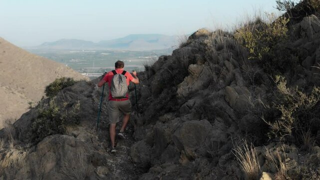 Off the beaten path, a man in a red shirt and shorts climbs steep rocks with trekking poles, seeking the summit in the morning sun.