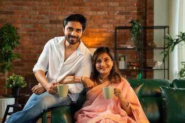 Elderly Indian mother and son sharing tea or coffee, enjoying casual conversation on the sofa