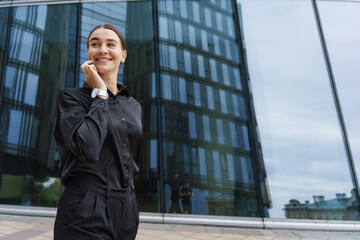 Young Professional Woman Smiles While Talking on Her Phone in Front of a Modern Glass Building on an Overcast Day