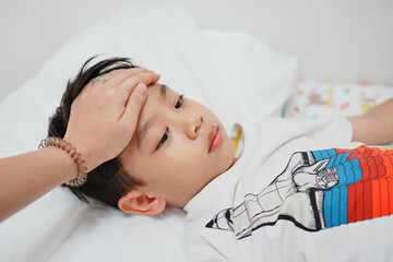 A young boy lies in bed while a hand checks his forehead for fever, showcasing care and concern in...