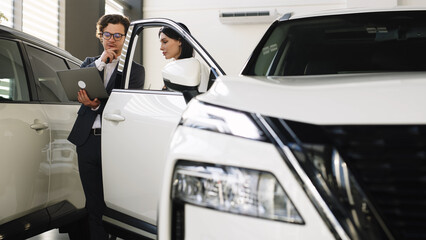 A fashionable young woman inspects a new car at a car dealership while being consulted by a manager. Concept of stylish car purchase or test drive.