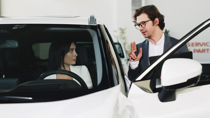 A fashionable young woman inspects a new car at a car dealership while being consulted by a manager. Concept of stylish car purchase or test drive.