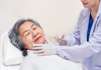 asian senior woman lying on couch in clinic,closed eyes,cosmetologist hands in gloves doing aesthetic procedure,rejuvenation injections for tightening and smoothing wrinkles on face skin